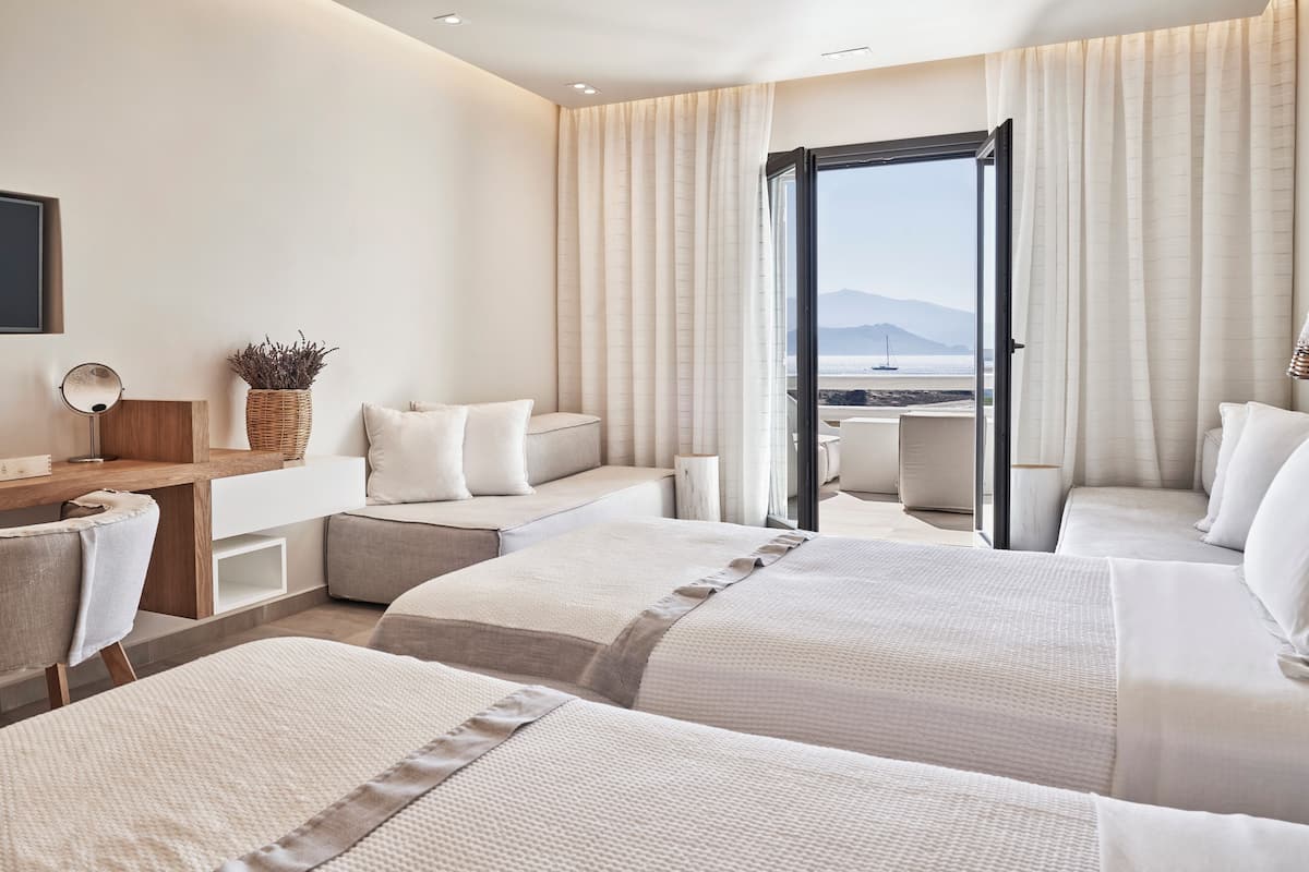 A modern hotel room with two beds, a desk, and a large window offering a view of the sea and mountains.