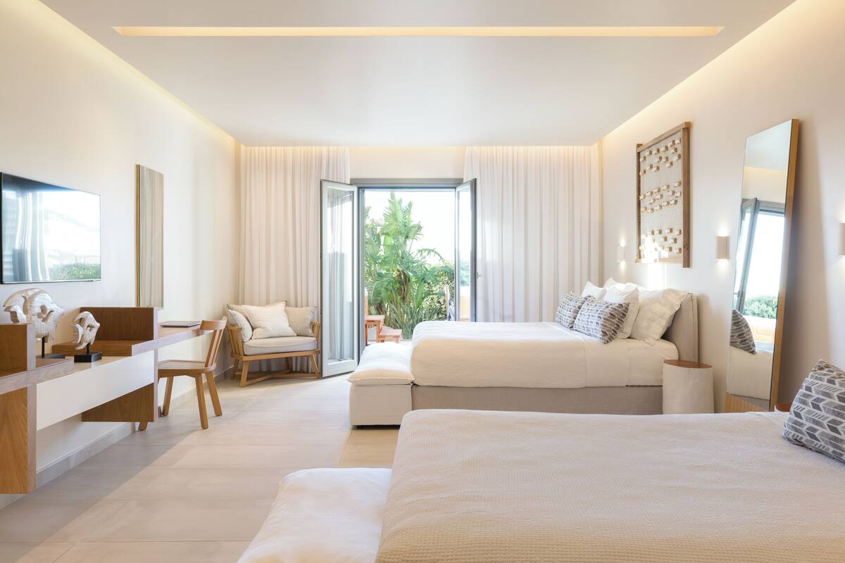 A modern bedroom with a large bed, a desk, and a chair. There is a window with curtains and a view of greenery outside.