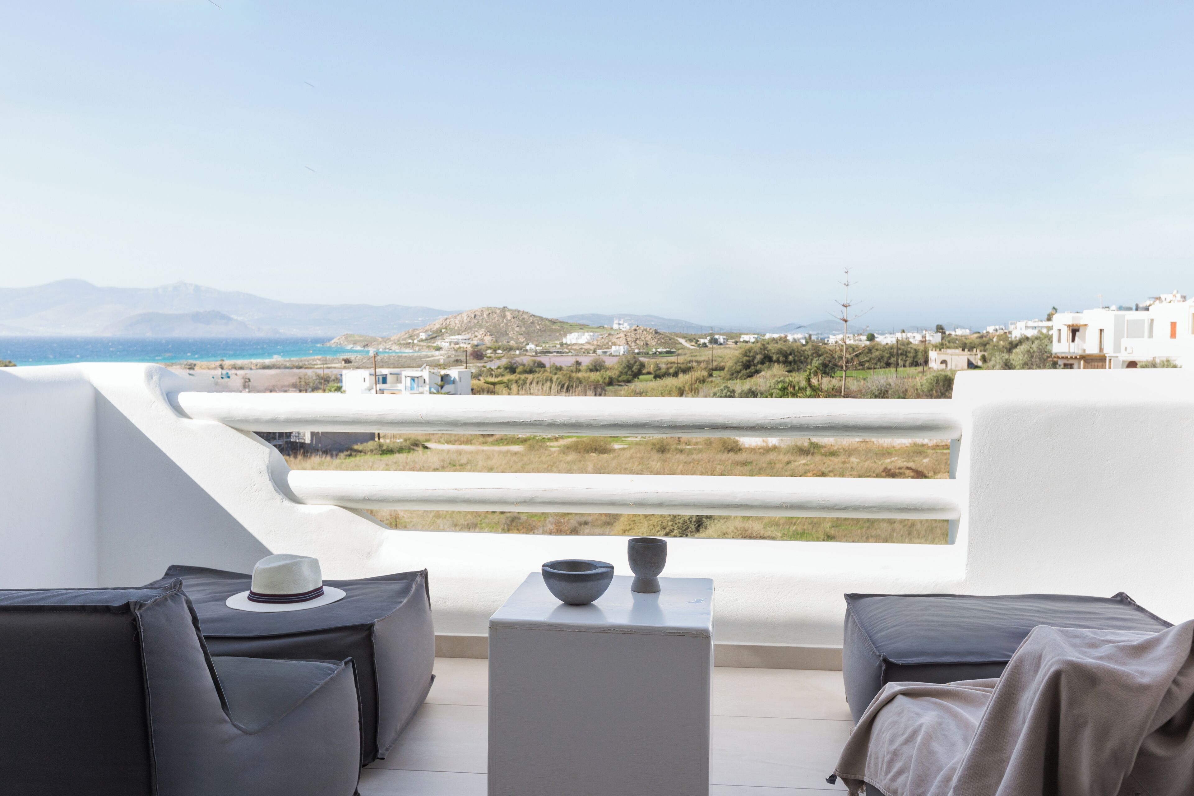 A balcony with a view of the sea and distant hills, featuring a sofa, a small table, and a hat.