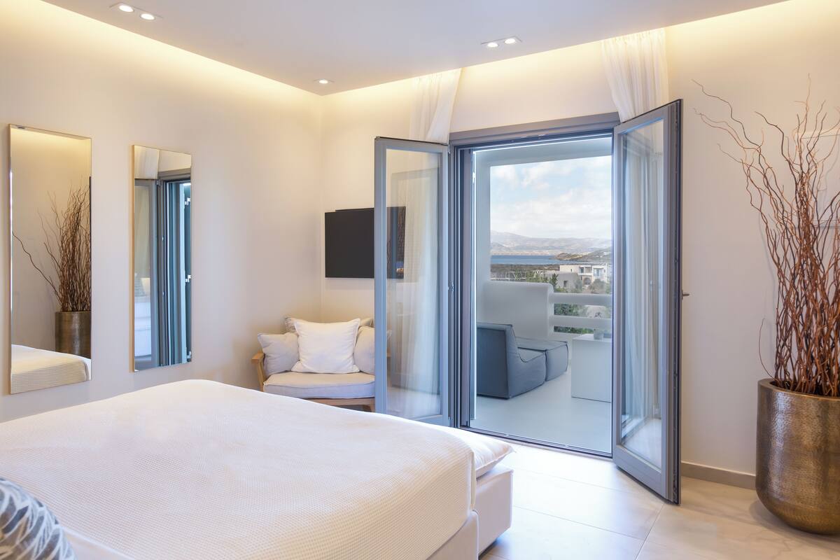 A modern bedroom with a large bed, a balcony with a view, and a TV mounted on the wall.
