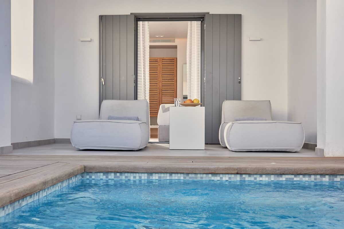 A poolside area with two white lounge chairs, a small table with a fruit bowl, and a wooden door leading to a room.