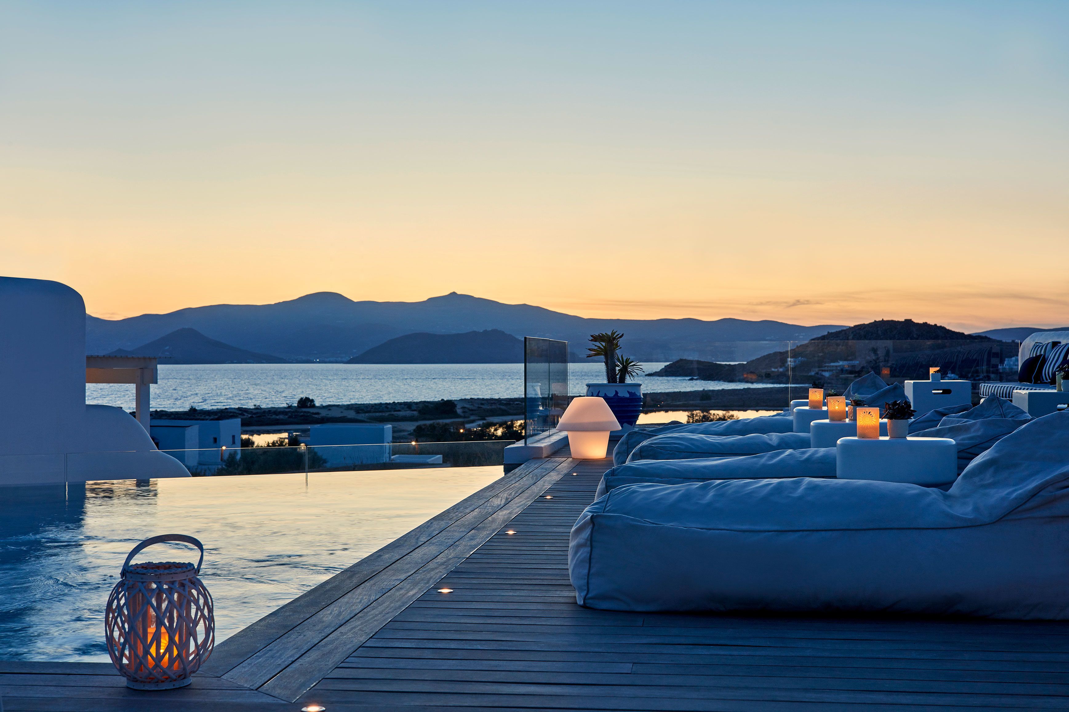 Un lounge en la azotea con piscina, asientos blancos acolchados y vistas al mar al atardecer.