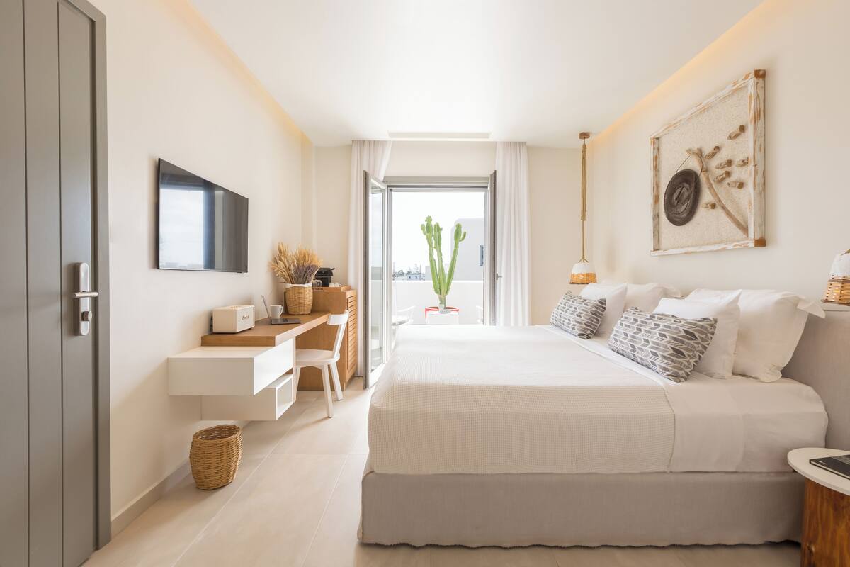 A modern bedroom with a bed, desk, and chair, a flat-screen TV, and a large window with a view.