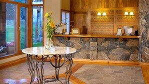 A spacious room with a wooden ceiling, a stone wall, and a large window. There is a round table with a vase of flowers and a chandelier above it. - Chamonix Posada y Spa (Villa General Belgrano)