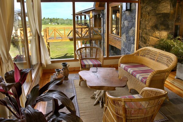 Una habitación acogedora con muebles de mimbre, una pared de piedra y vistas a una terraza de madera y follaje exterior.