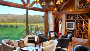 A cozy interior with a wooden ceiling, large windows, and a view of a green landscape. There are leather chairs, a television, and a wine rack. - Chamonix Posada y Spa (Villa General Belgrano)