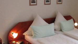 A hotel room with a wooden headboard, two beds with white bedding and light green pillows, a bedside lamp, a small table with a lamp and a remote control, and three framed pictures on the wall. - Neuenkirchener Hof (Neuenkirchen)