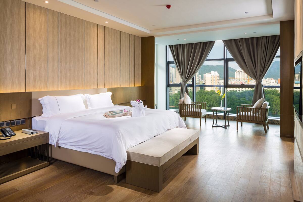 Une chambre d’hôtel moderne dotée d’un grand lit, de tables de chevet, d’un banc, d’une petite table avec des chaises et d’une grande fenêtre donnant sur la ville.