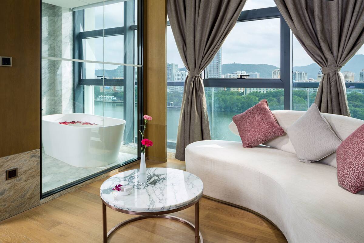 Une chambre d’hôtel moderne avec une grande fenêtre, une baignoire blanche, une table ronde en marbre et un canapé agrémenté de coussins à motifs.