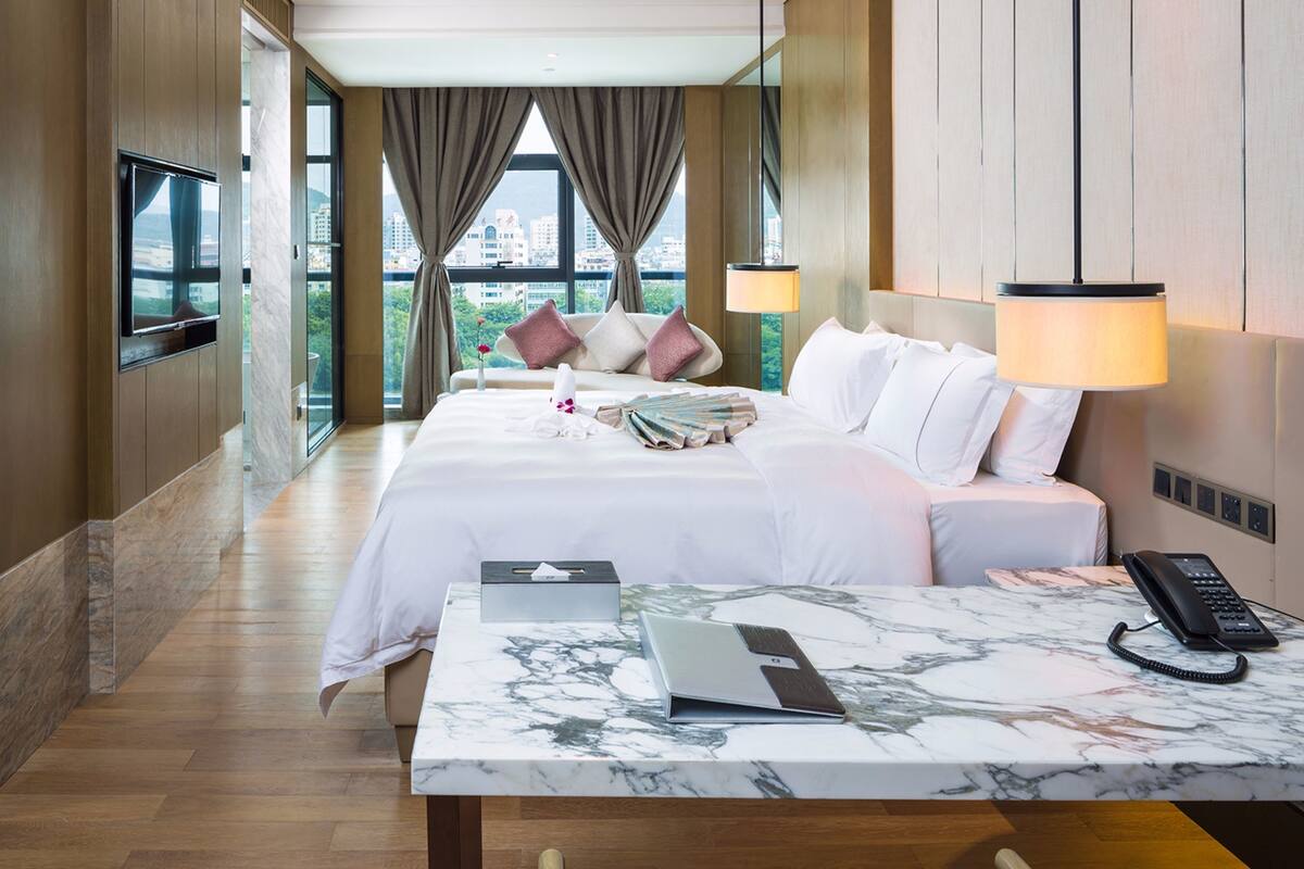 Une chambre d’hôtel avec un grand lit, un bureau avec un ordinateur portable, un téléphone et une vue sur la ville grâce à de grandes fenêtres.