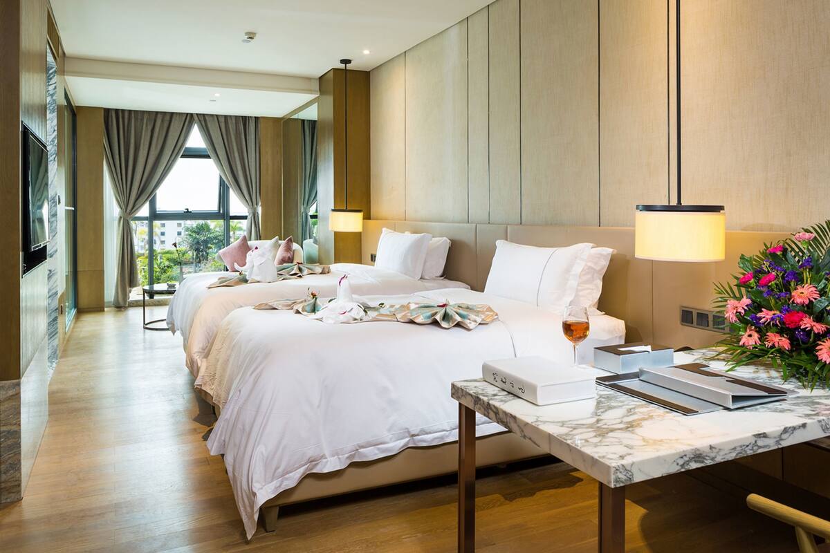 Une chambre d’hôtel avec un grand lit, un bureau avec un ordinateur portable, un vase de fleurs et une fenêtre avec des rideaux.