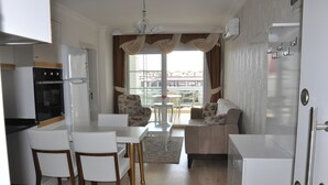 A compact kitchen with white appliances, a dining area with white chairs, and a sofa. - Akbük Palace Hotel & Residence (Didim)