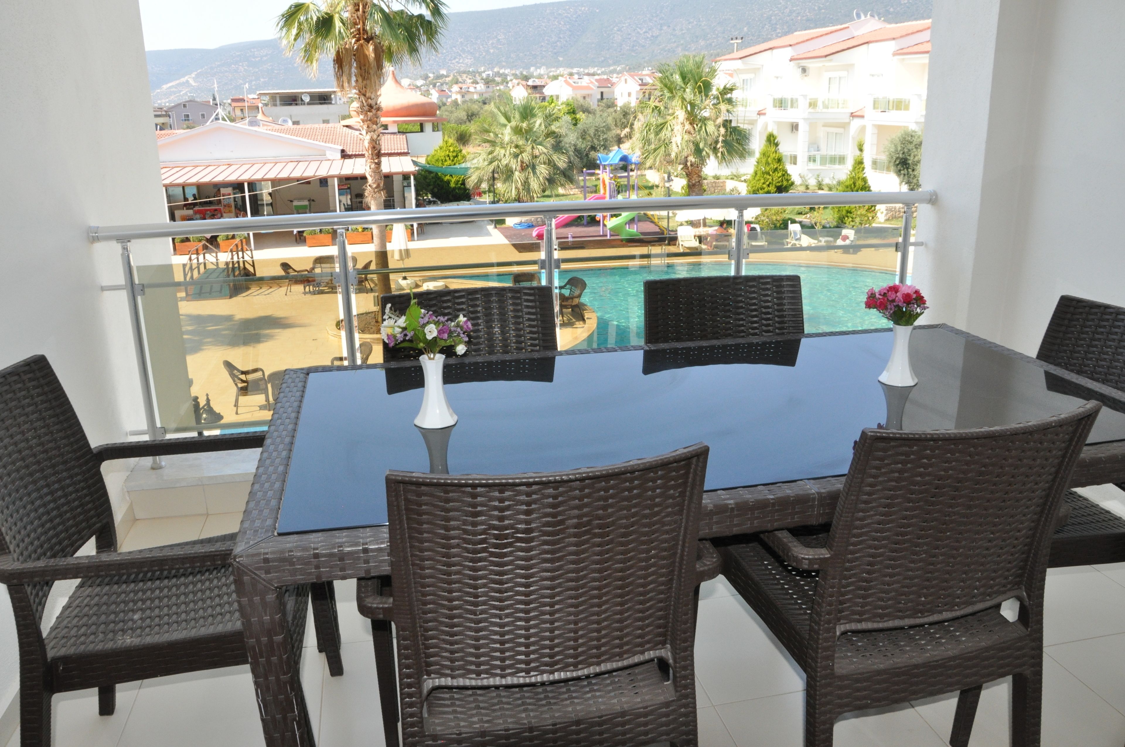 A balcony with a glass table and wicker chairs, overlooking a swimming pool and a resort area.