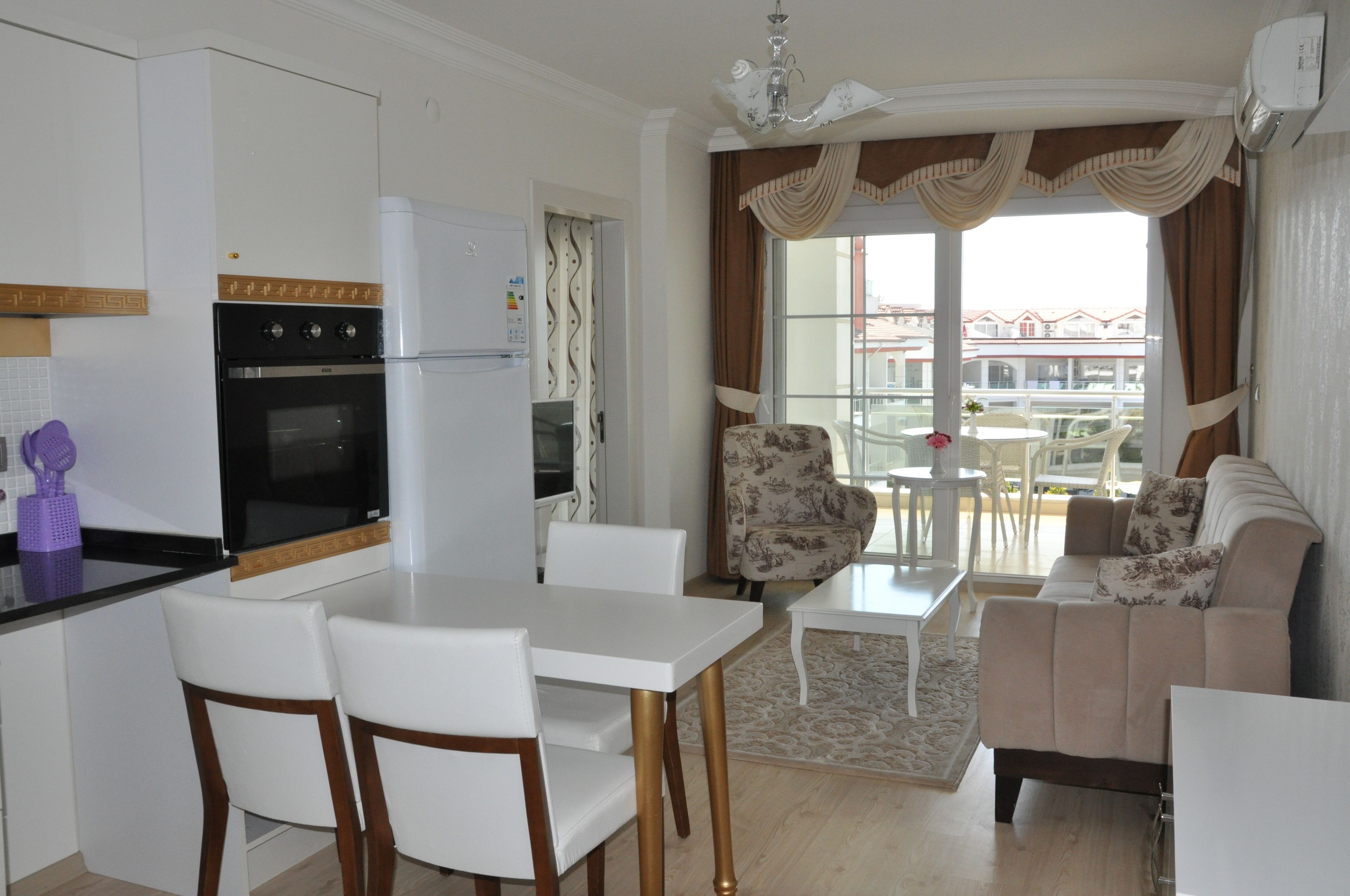 Une cuisine moderne avec des meubles blancs, un four noir et un réfrigérateur blanc. On y trouve un coin repas avec une table et des chaises blanches, ainsi qu’un canapé recouvert d’un tissu à motifs floraux.