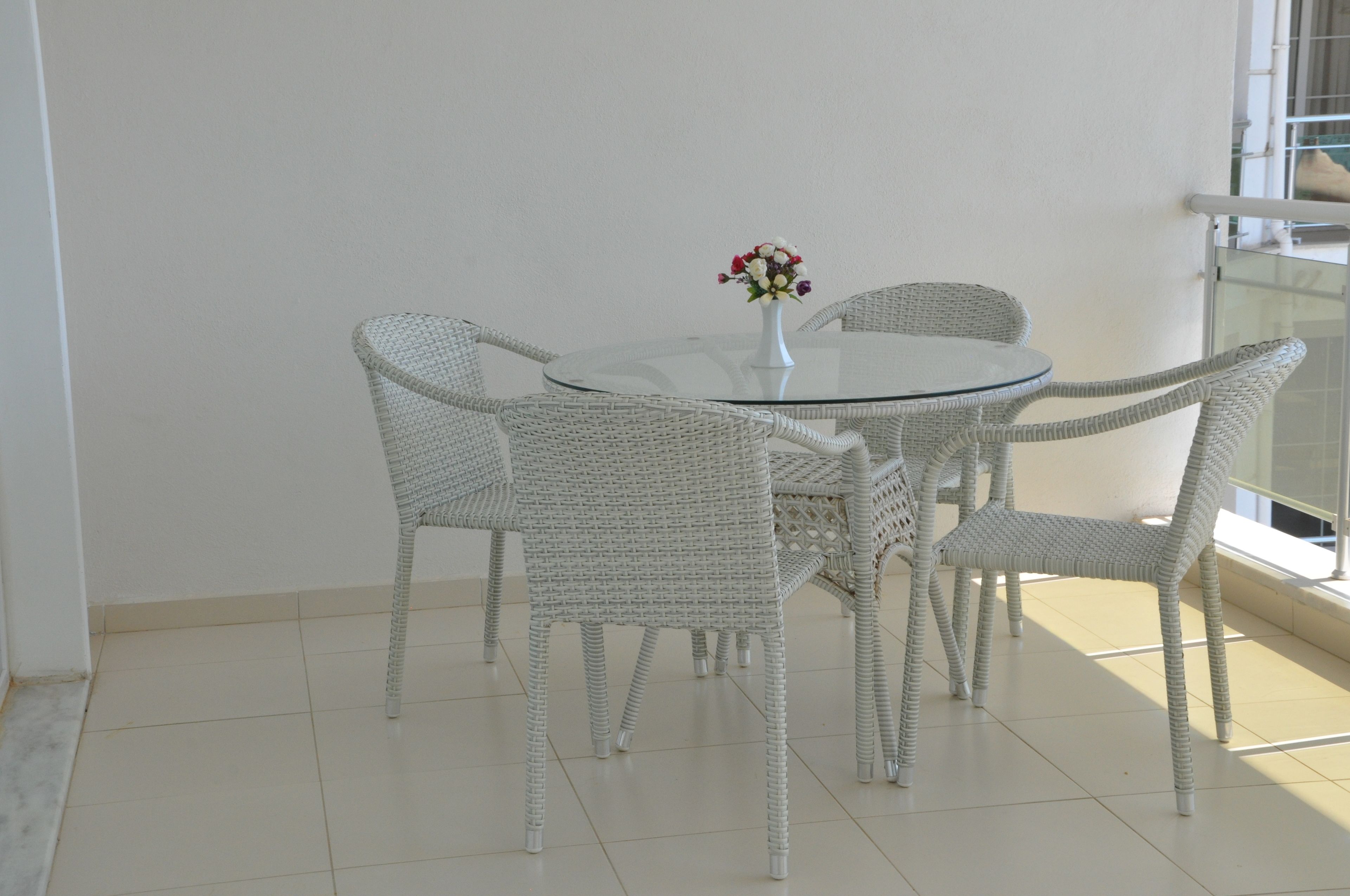 A round glass table with four wicker chairs, a white vase with flowers, and a balcony with a railing.