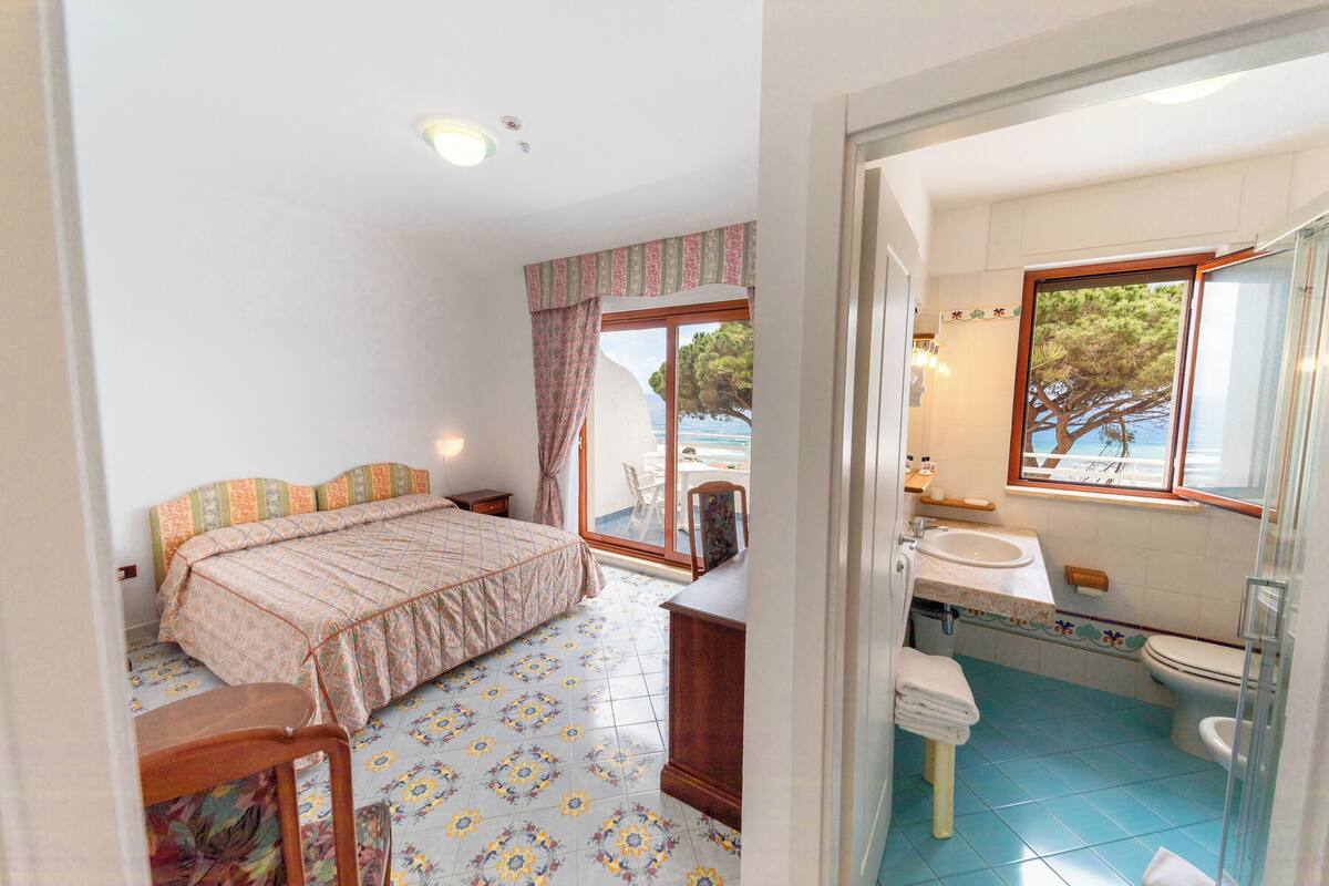 A bedroom with a bed, a chair, a desk, and a view of the sea through a window.
