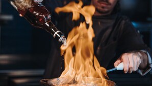 A chef cooking with a pan on a stove, with flames visible. - Revier Mountain Lodge Lenzerheide (Vaz-Obervaz)