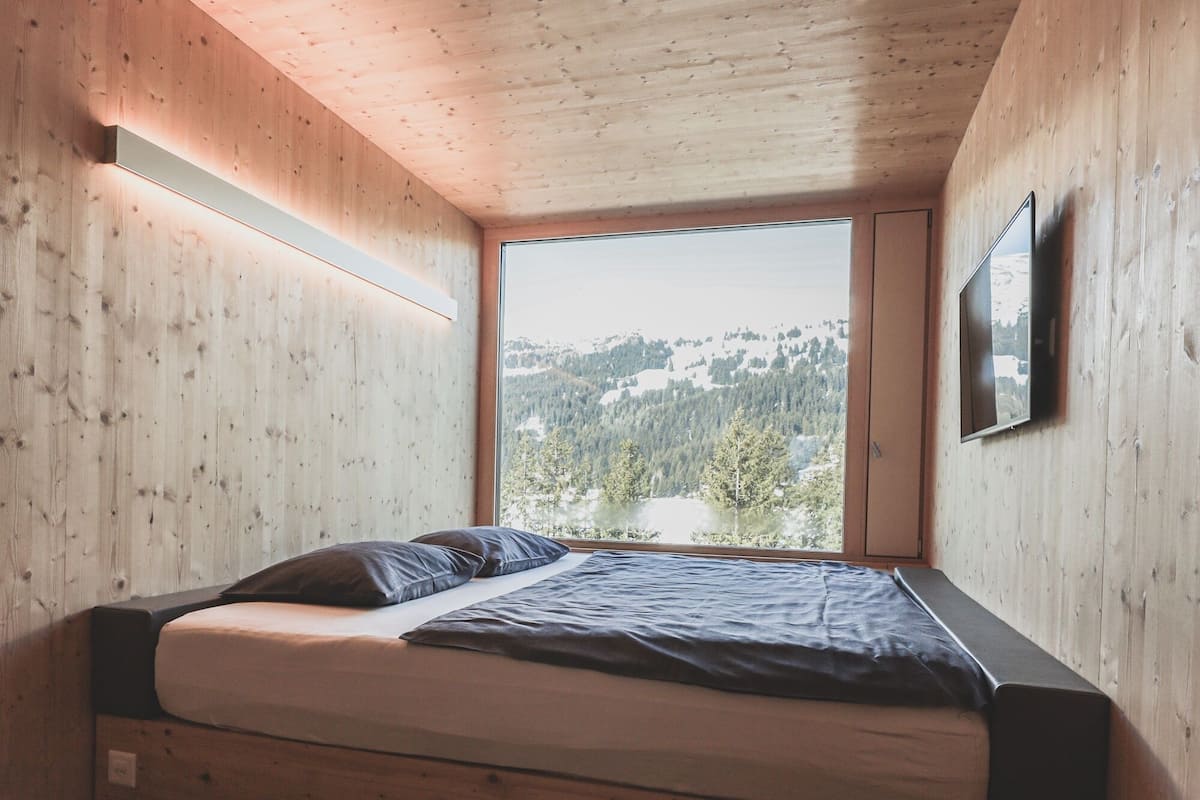 A bedroom with a wooden ceiling, a bed with a mattress and pillows, a window with a view of a forest and mountains, and a mounted television.