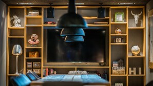 A modern living room with a large flat-screen TV, wooden shelving with various decorative items, a dining table with chairs, and a hanging pendant light. - Revier Mountain Lodge Lenzerheide (Vaz-Obervaz)