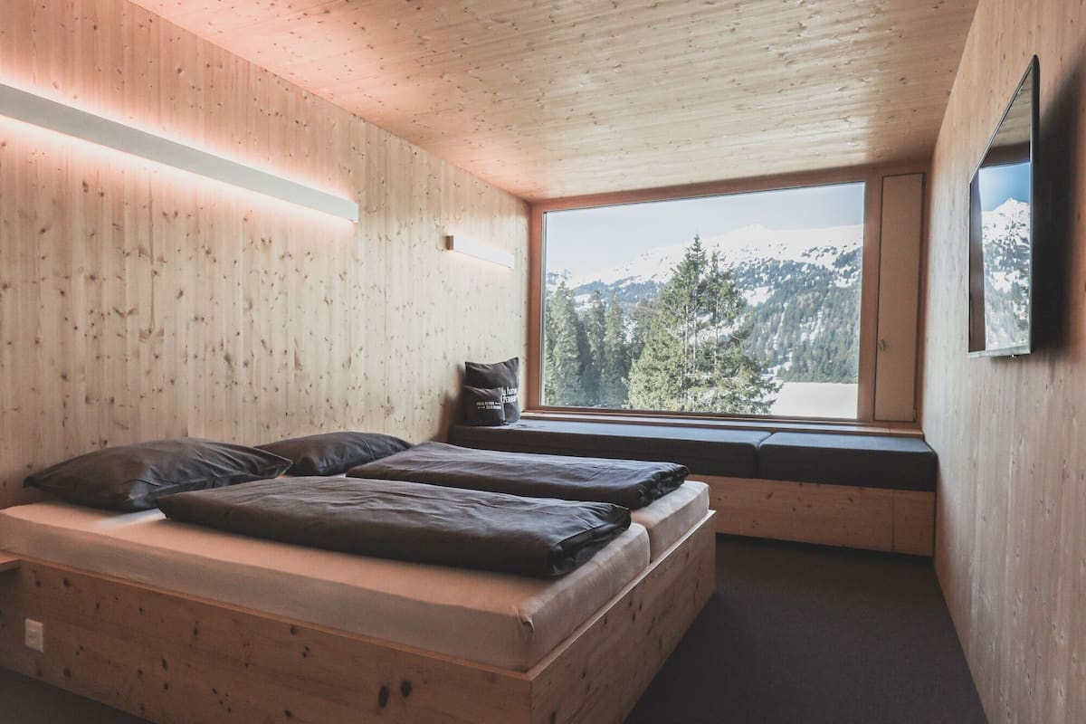 A wooden cabin interior with a bed, a window with a mountain view, and a mounted picture on the wall.