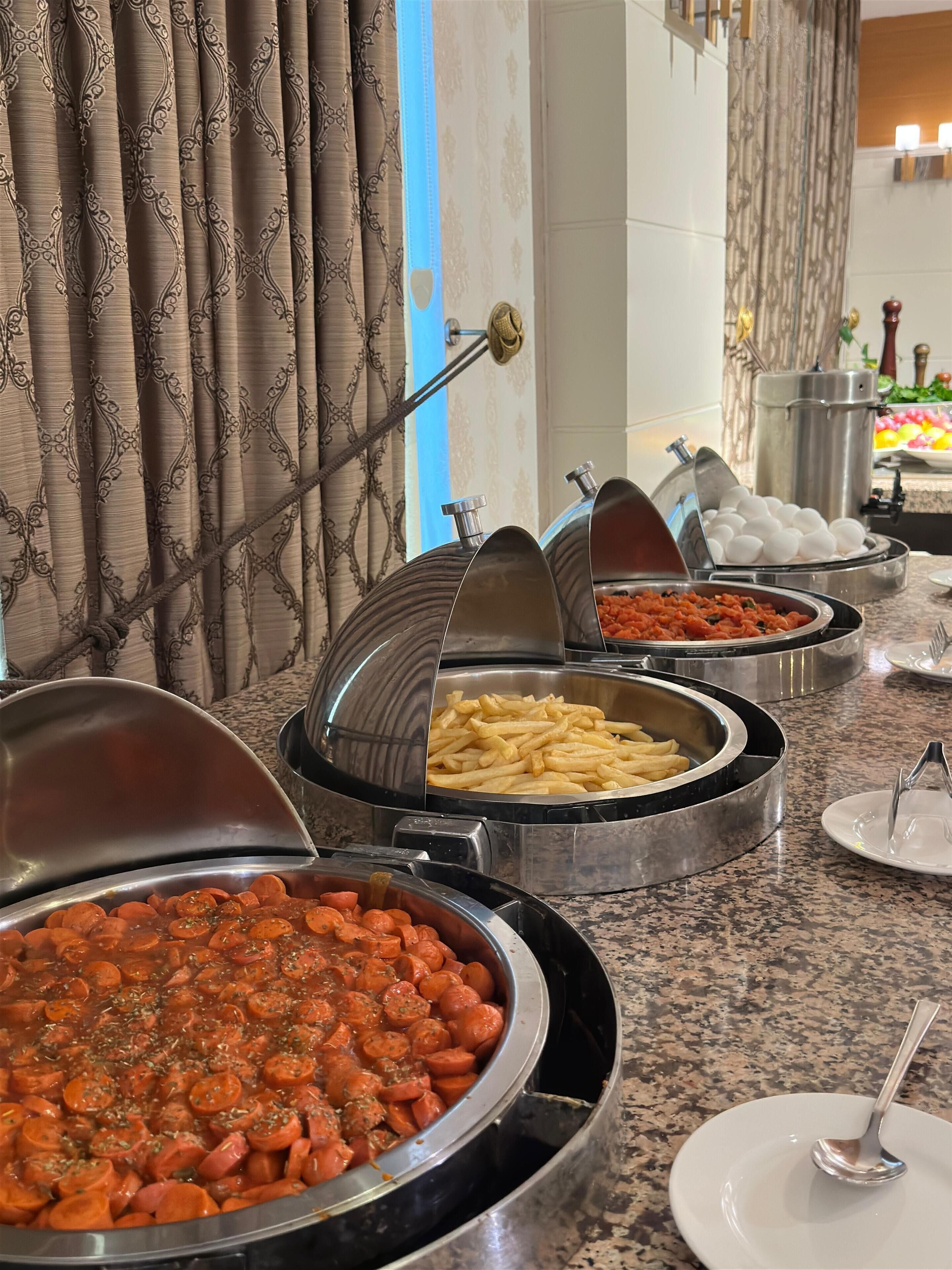 A buffet with various food options, including a large container of sliced sausages, a bowl of French fries, and a dish of mixed vegetables.