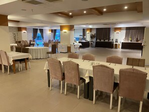 Un comedor espacioso con varias mesas y sillas, un área de cocina al fondo, y ventanas grandes con cortinas.
