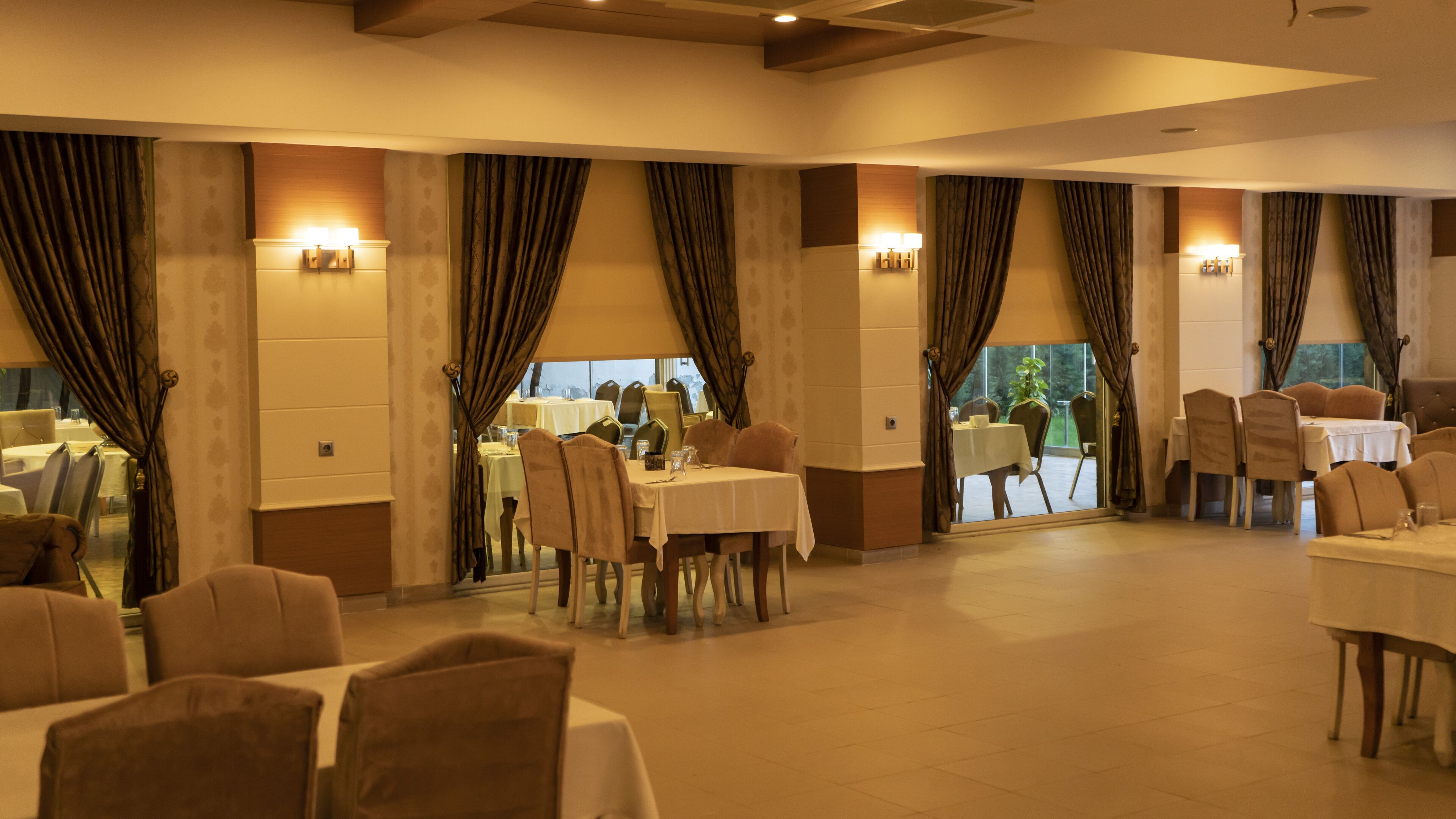 A spacious dining area with multiple tables and chairs, large windows with curtains, and wall-mounted lighting.