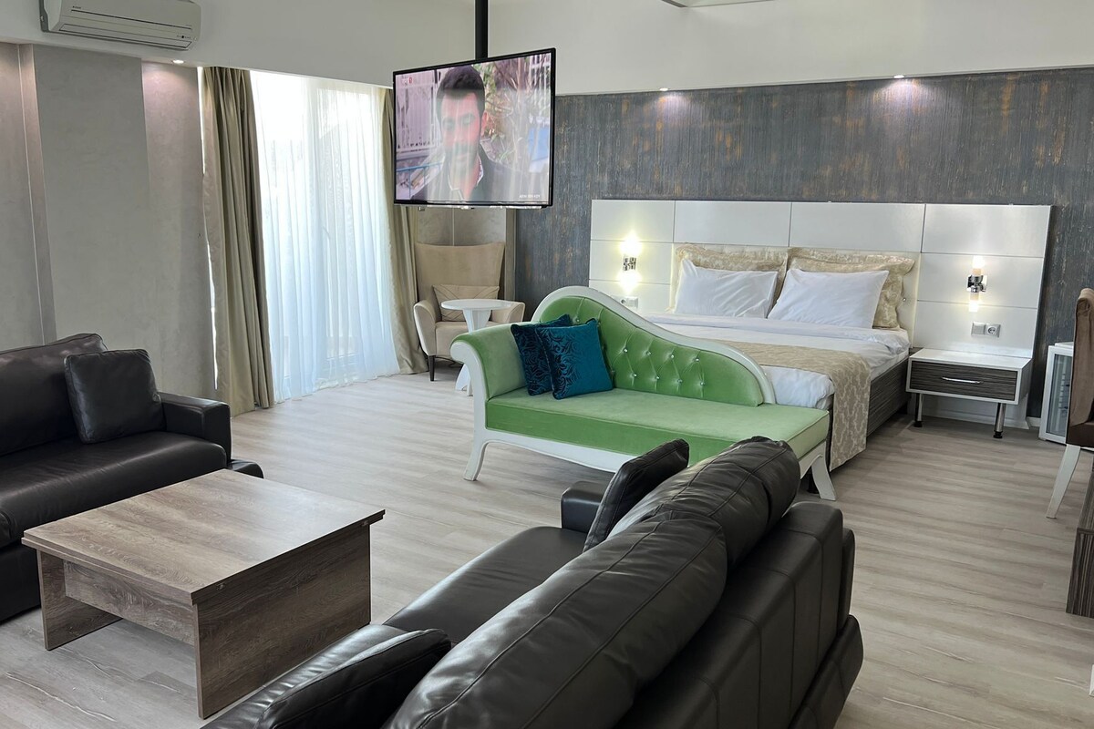 Una habitación de hotel moderna con un sofá verde, sofás de cuero, una mesa de centro de madera, una cama con ropa blanca y un televisor montado en la pared.