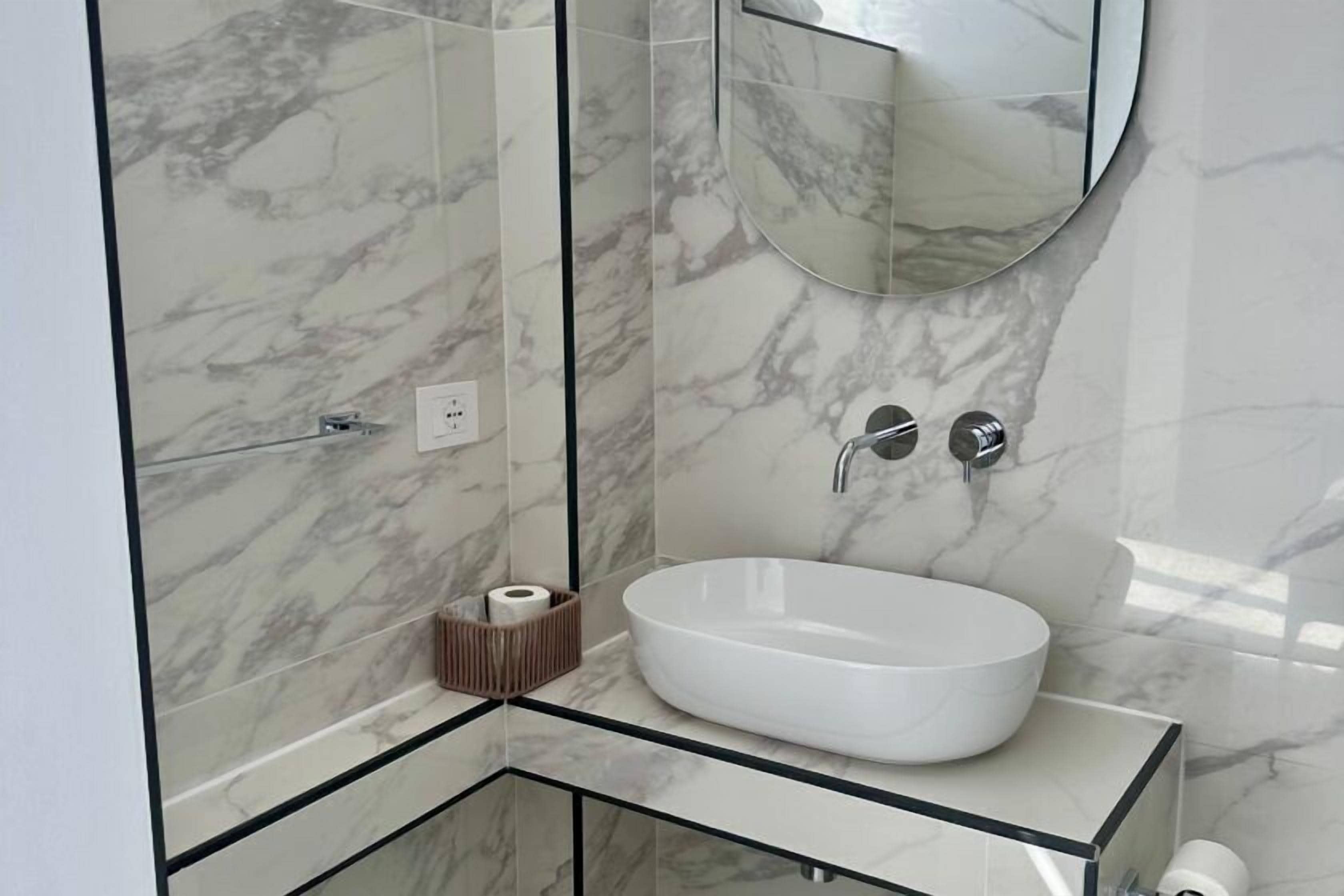 A modern bathroom with a marble backsplash, a round white sink, and a mirror above it.