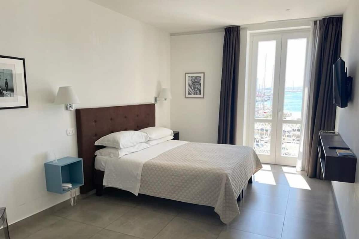 A hotel room with a bed, a small blue side table, a wall-mounted TV, and a balcony with a view of the sea.