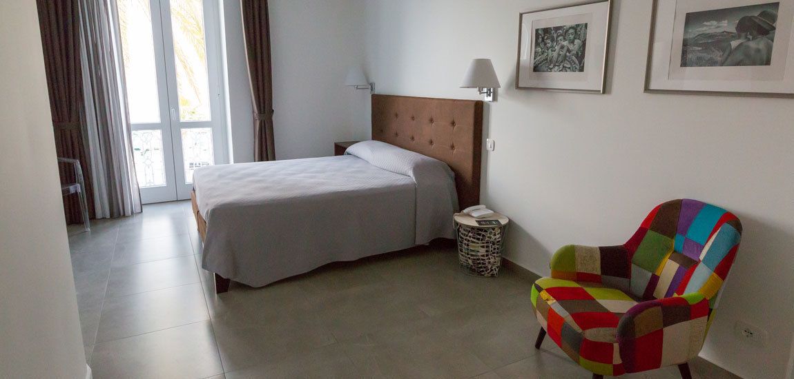 Quarto de hotel com cama, uma cadeira, mesa de cabeceira e luminária.