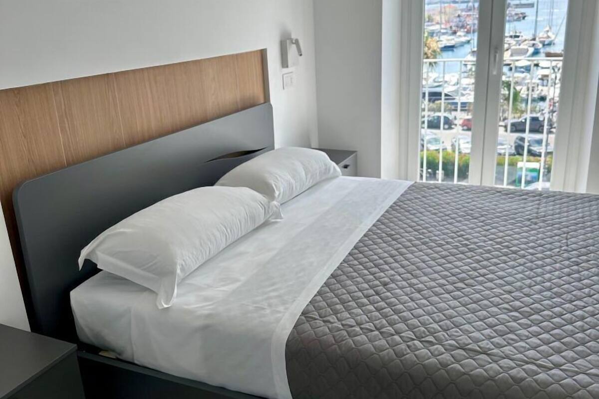 A neatly made bed with white linens and a grey quilted cover, a wooden headboard, and a nightstand with a lamp.
