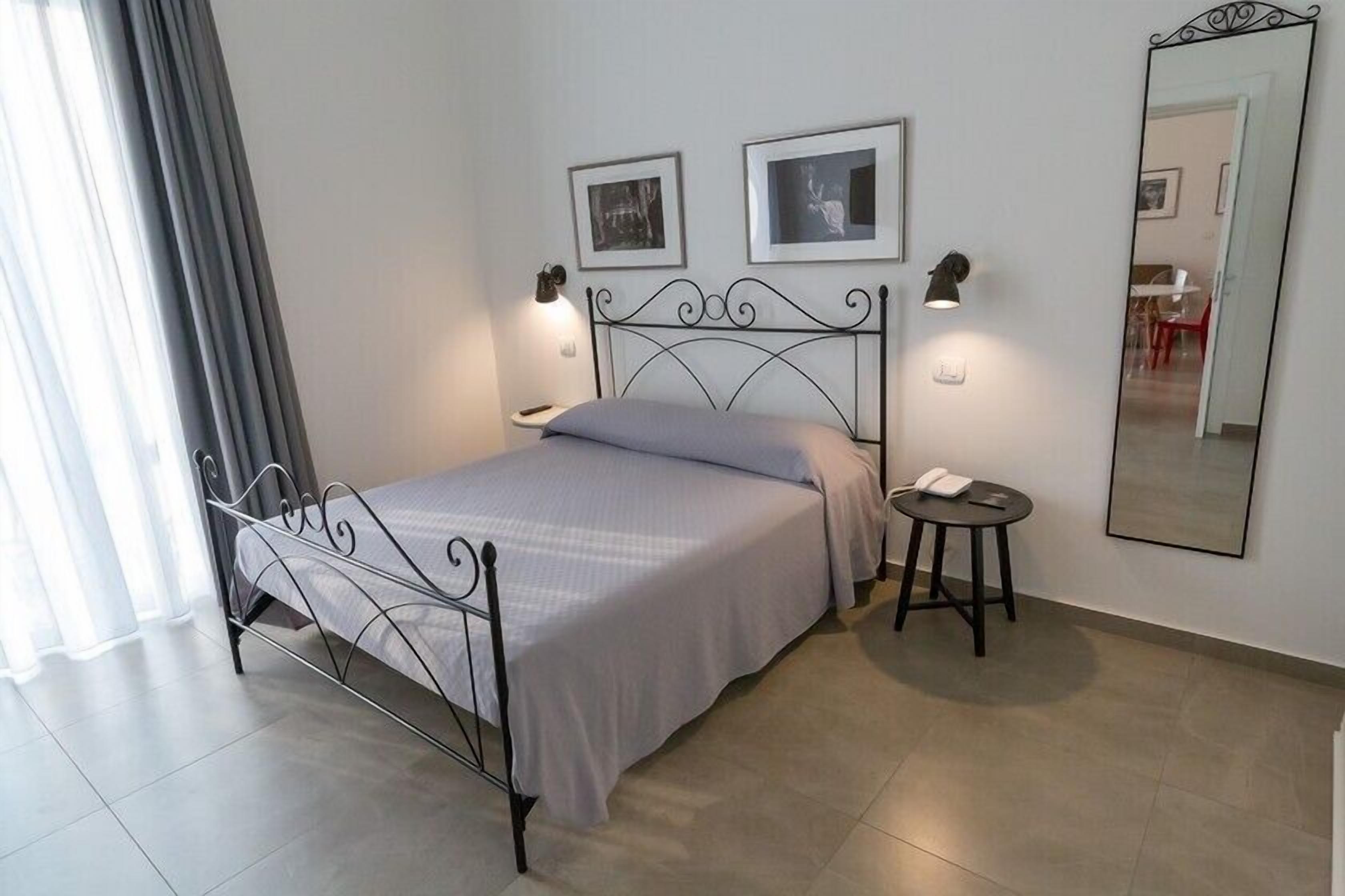 Un dormitorio ordenado con estructura de cama metálica, mesita de noche con lámpara y espejo en la pared.