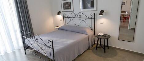 Un dormitorio ordenado con estructura de cama metálica, mesita de noche con lámpara y espejo en la pared.