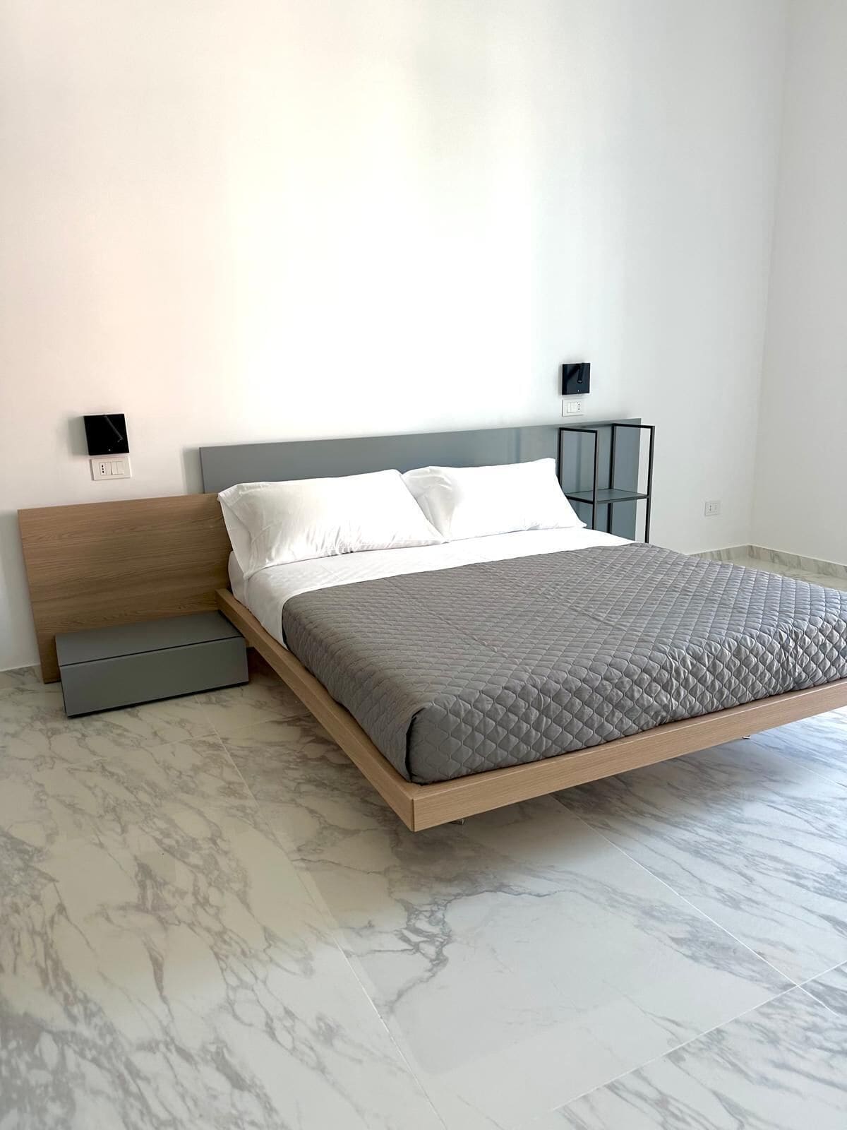Quarto moderno com design minimalista, apresentando uma cama com colchão acolchoado cinza, cabeceira de madeira e móvel de armazenamento cinza sob a cama.
