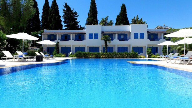 Une piscine avec des chaises longues et des parasols, un bâtiment blanc aux volets bleus, et de grands arbres en arrière-plan.