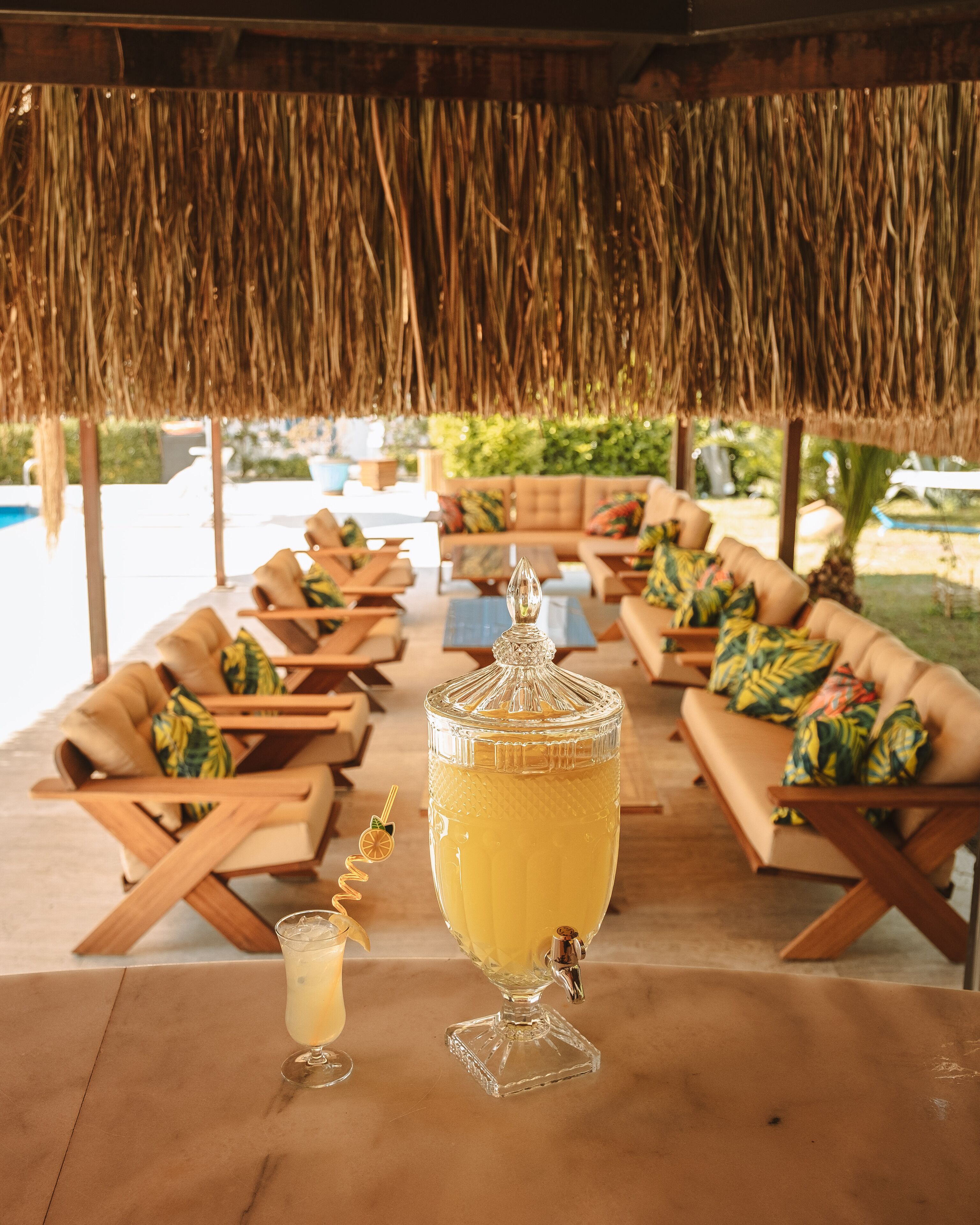 A tropical outdoor seating area with thatched roofs, cushioned lounge chairs, and a large beverage dispenser.