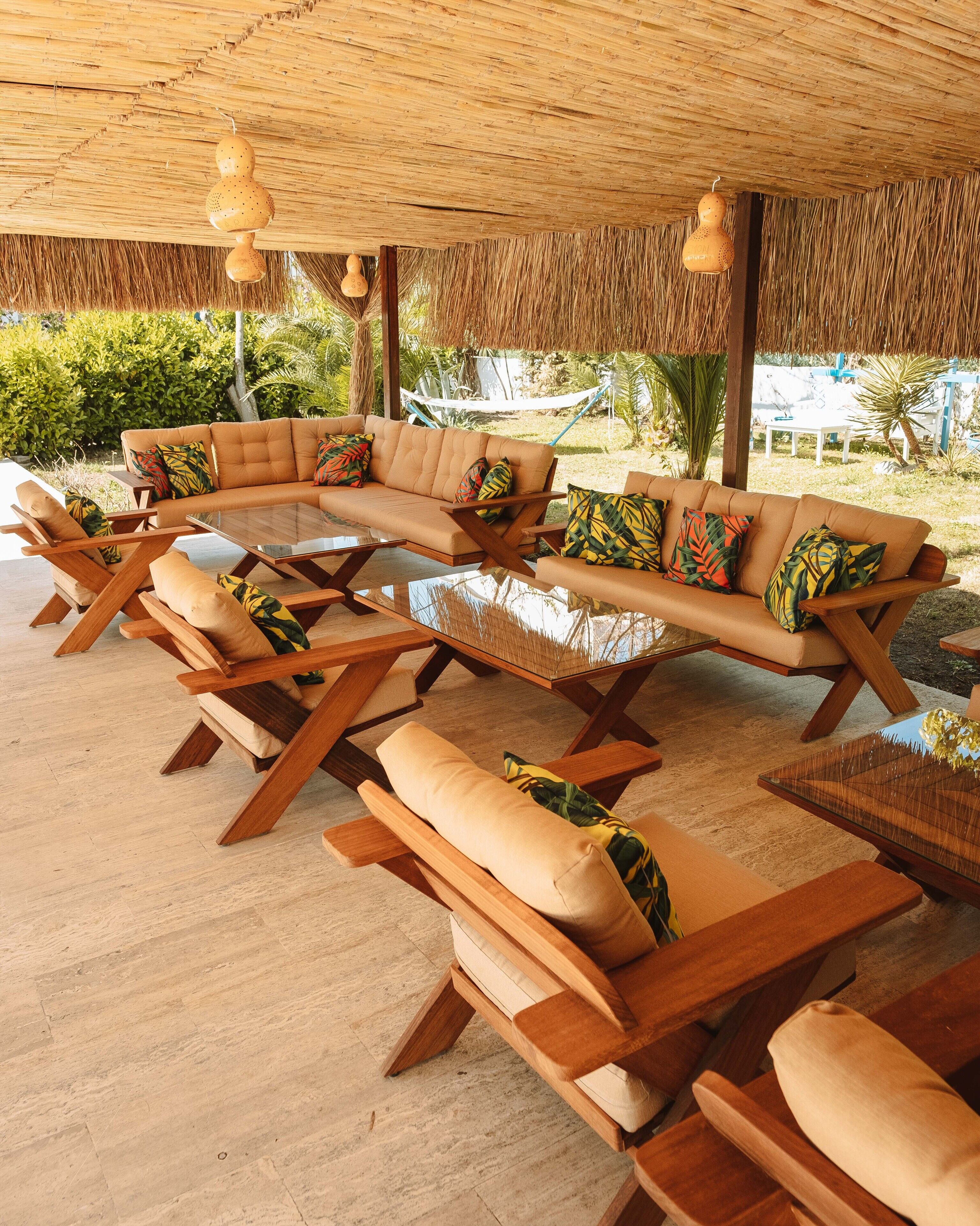 A sunlit outdoor seating area with a thatched roof, cushioned sofas, wooden tables, and hanging light fixtures.