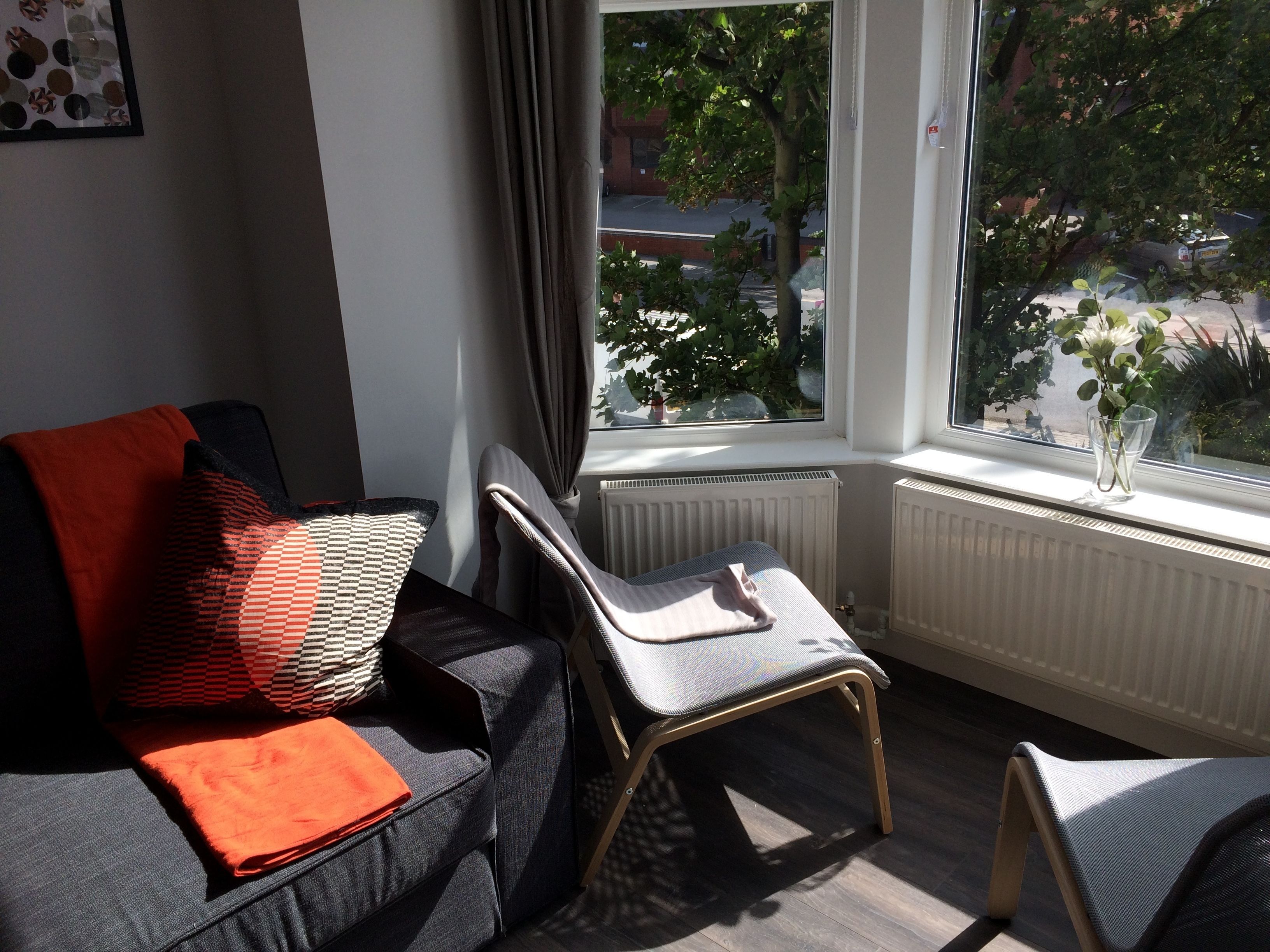 A modern living room with a grey sofa, a small wooden chair, and a window with a view of the street and trees.