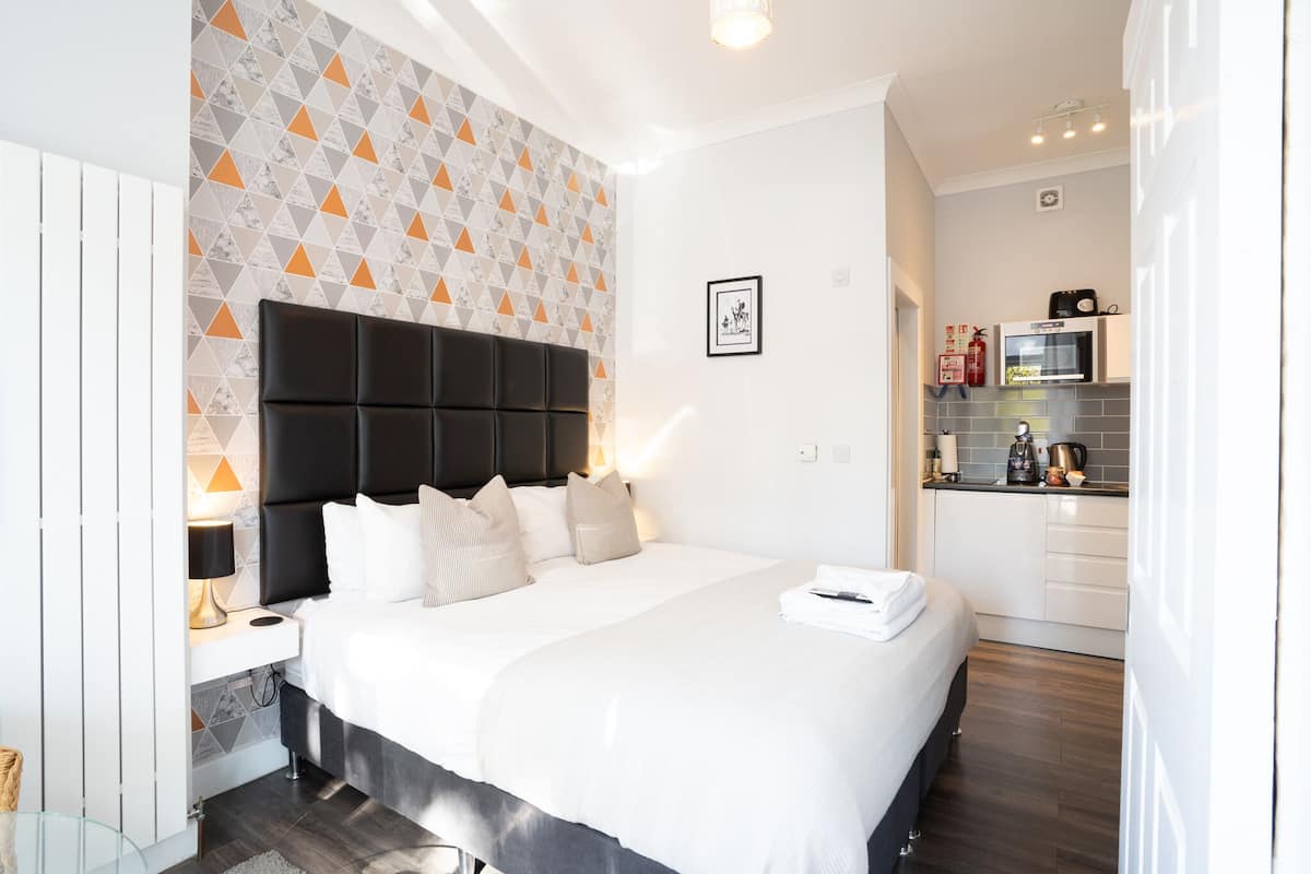 Ein modernes Hotelzimmer mit einem großen Bett, einer festen Kopfteil, einem Nachttisch mit Lampe und Blick in den Küchenbereich.