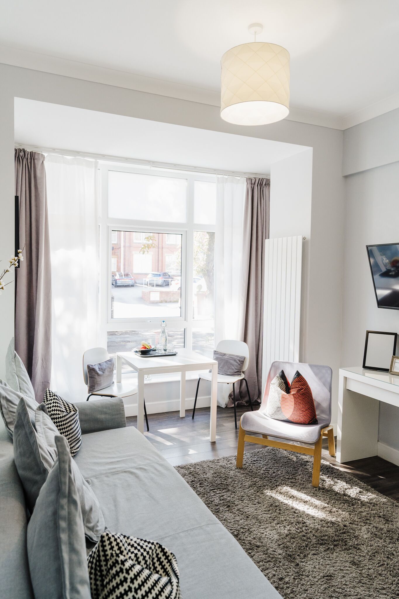 Un salon moderne avec un canapé, une table basse et un fauteuil. Il y a une fenêtre avec des rideaux, une lampe et une vue sur la rue.