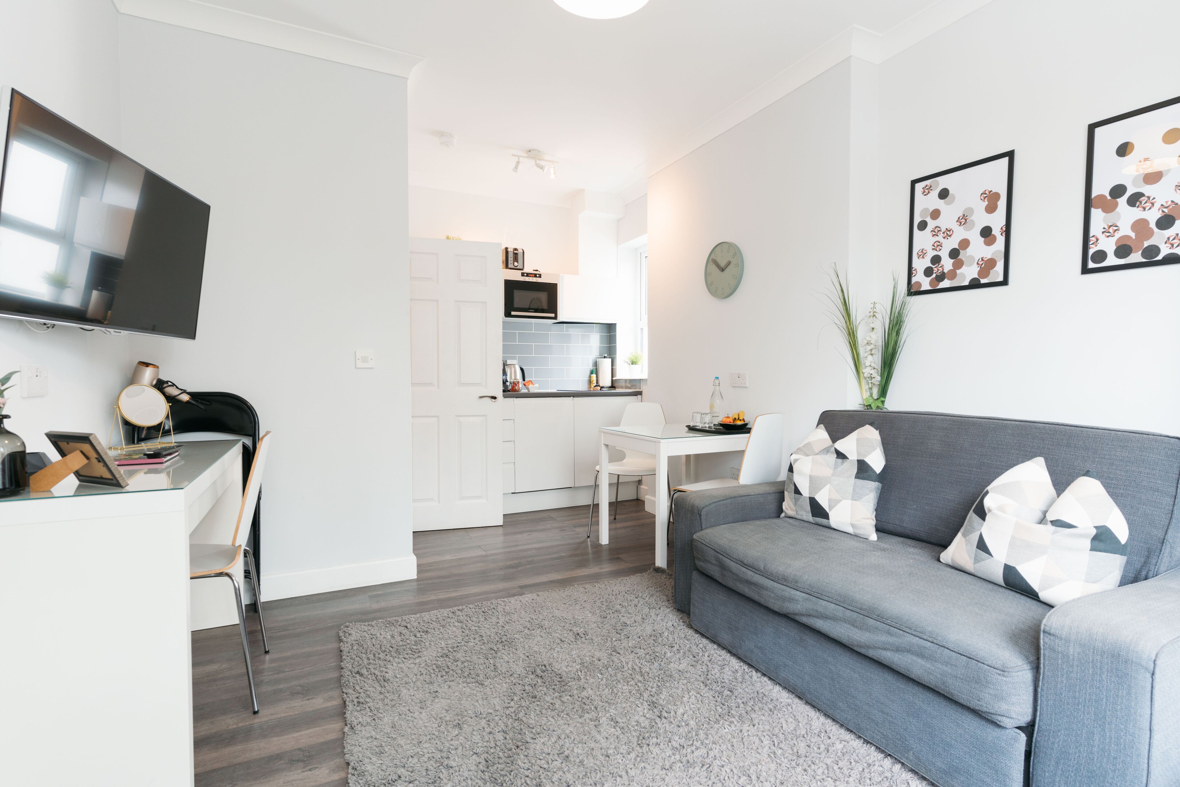A modern living room with a grey sofa, a white dining table, and a kitchen in the background.