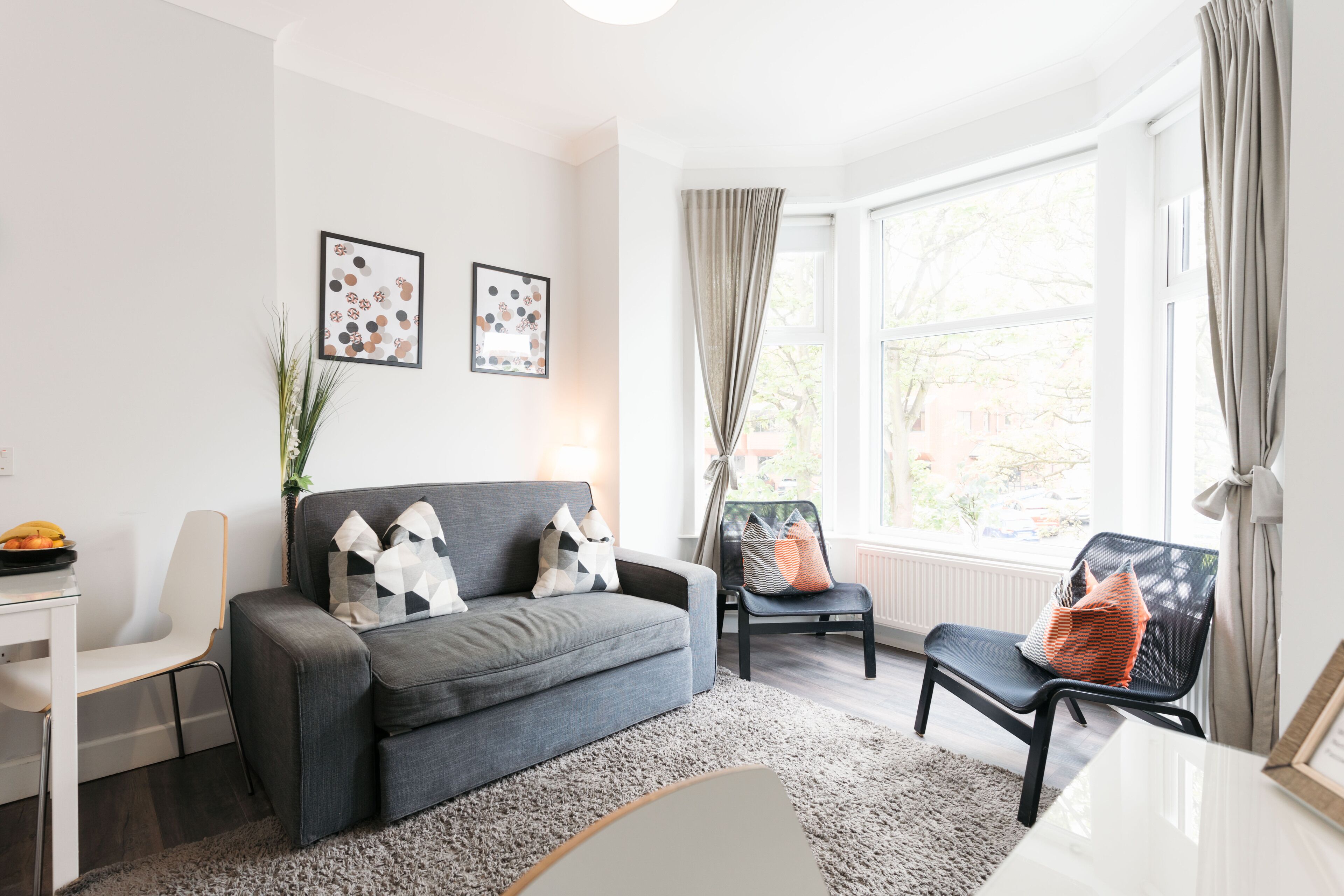 A modern living room with a grey sofa, a black chair, a white desk, and a large window with curtains.