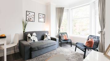 A modern living room with a grey sofa, a black chair, a white desk, and a large window with curtains.