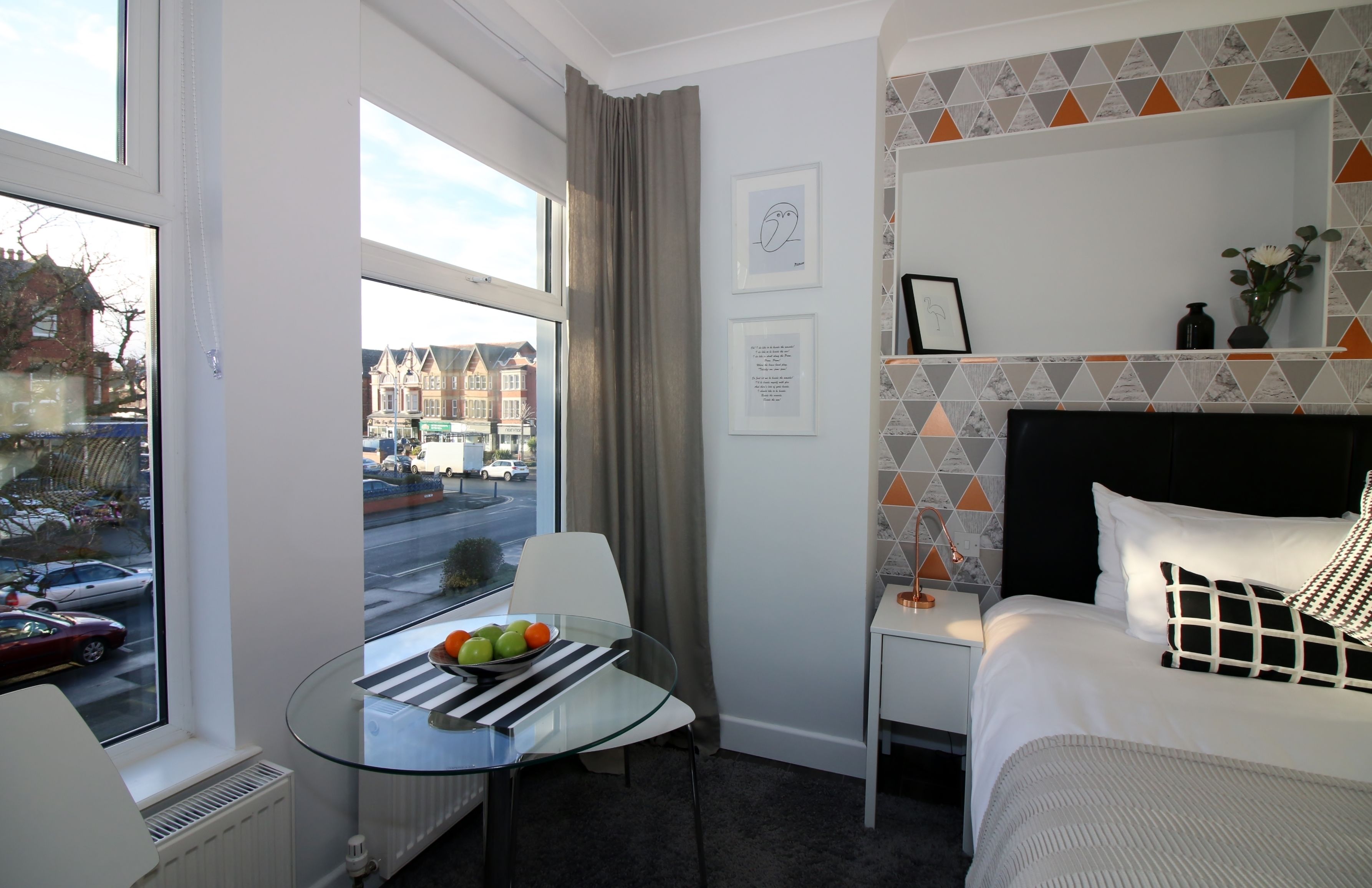 Une chambre d’hôtel avec un lit, une petite table sur laquelle se trouvent des fruits, une chaise, une fenêtre donnant sur la rue et une étagère avec des photos encadrées.
