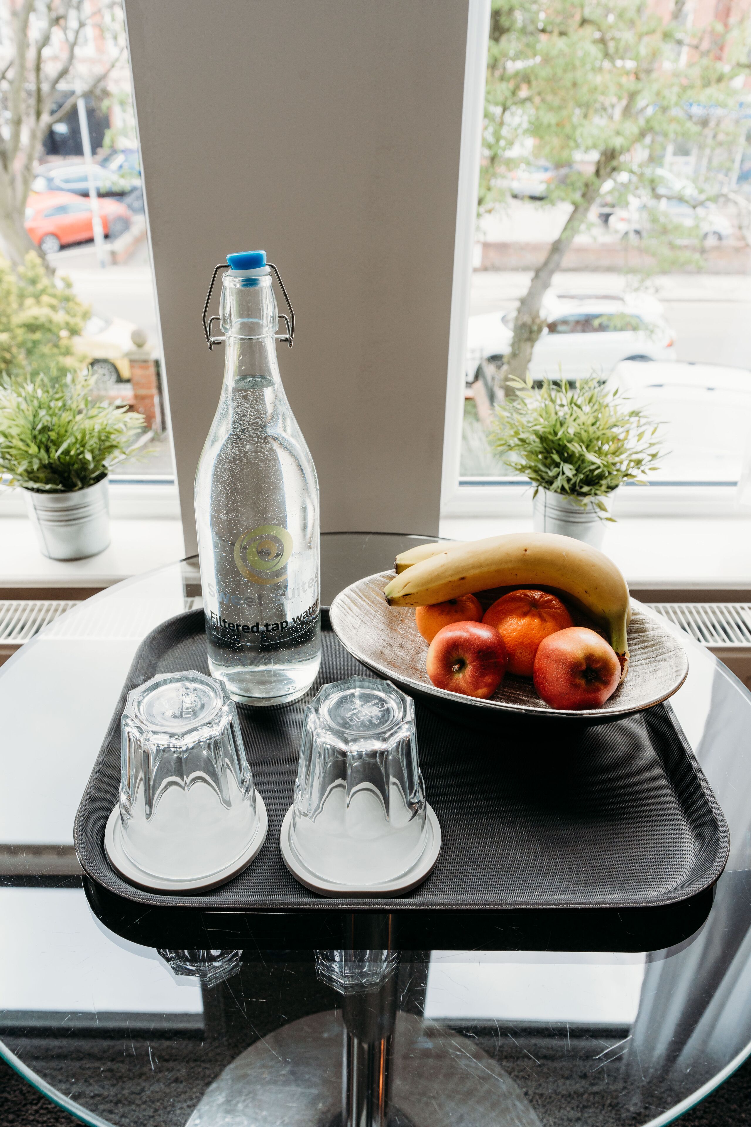 A clear glass bottle of filtered tap water, a banana, and apples on a tray by a window with potted plants.