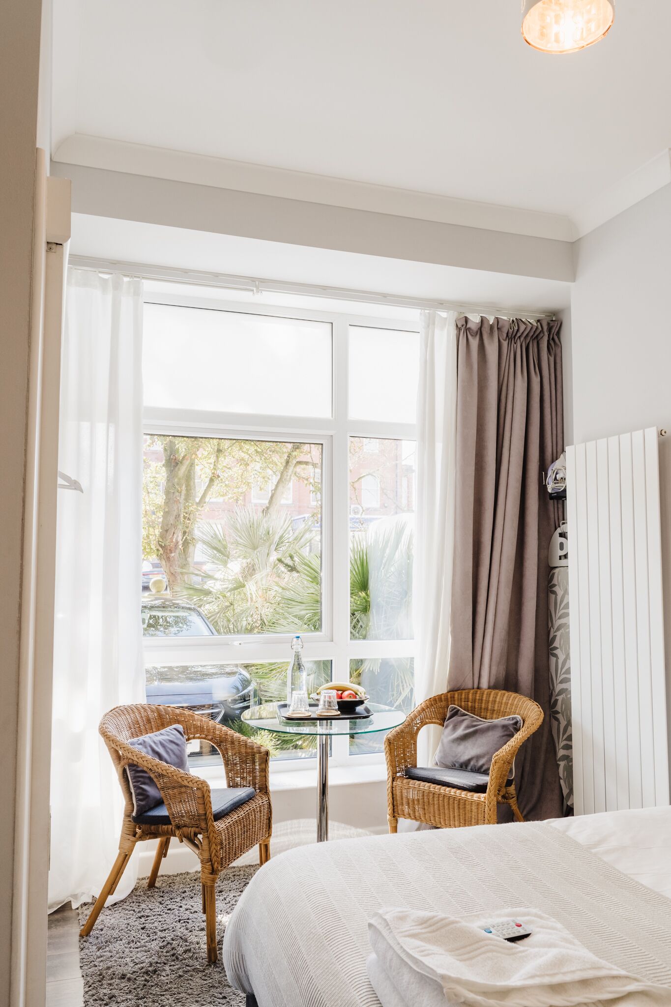 Une chambre avec une grande fenêtre, deux fauteuils en osier, un lit et une petite table sur laquelle se trouvent un vase et des fruits.