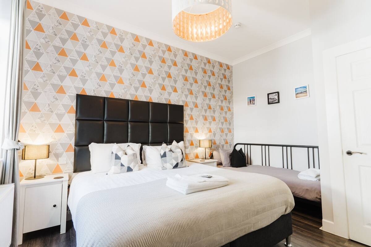 Ein modernes Hotelzimmer mit einem großen Bett, einem Kopfteil mit geometrischem Muster, zwei Nachttischen mit Lampen und einer Tür.