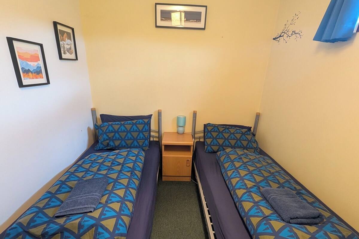 Ein kleines Zimmer mit zwei Einzelbetten, einem Nachttisch, einem Spiegel und einem Fenster mit Vorhängen.