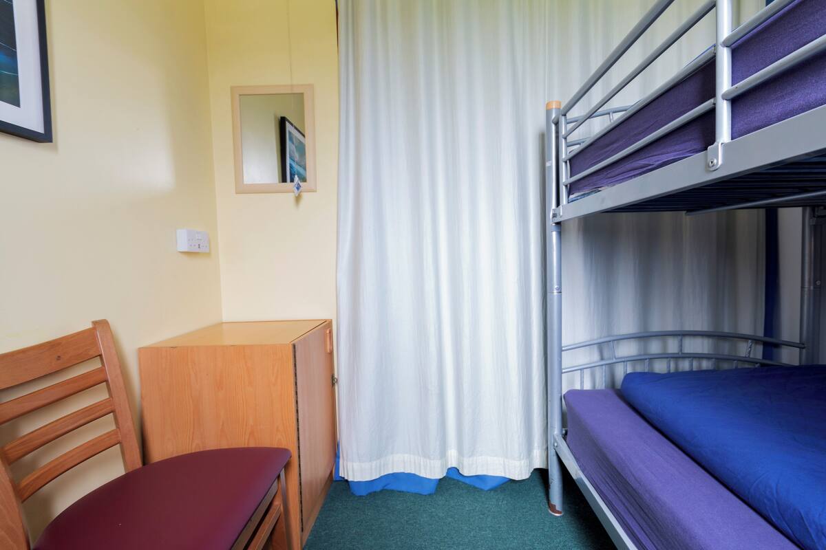 Ein Etagenbett mit einer violetten Matratze, ein Holztisch, ein Stuhl und ein weißer Vorhang.